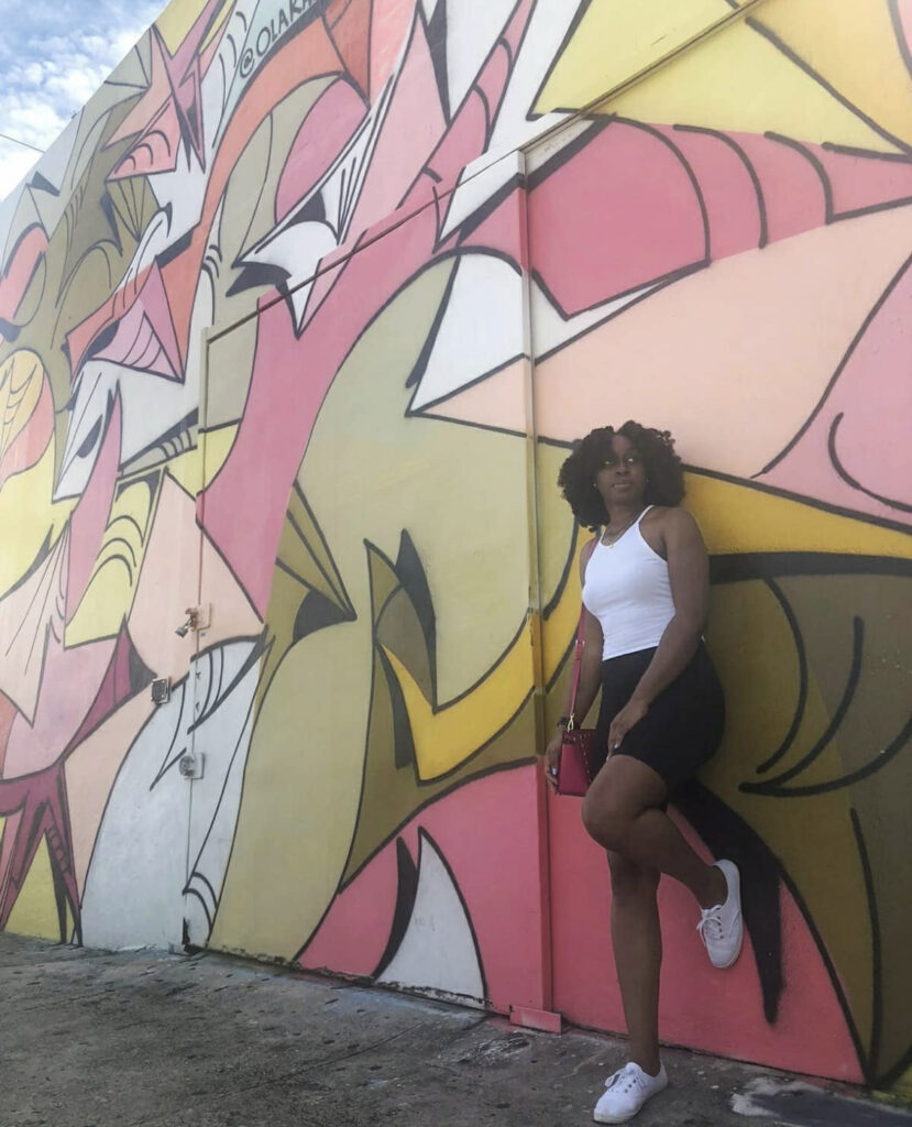 Woman On A Weekend Trip To Miami. Woman Leaning Against An Art Wall Mural In Miami, Florida. Mural Has Pink, Yellow, Brown And White Colors. Woman Traveling The World. Immersive Experiences In Miami, Florida. Travel USA.